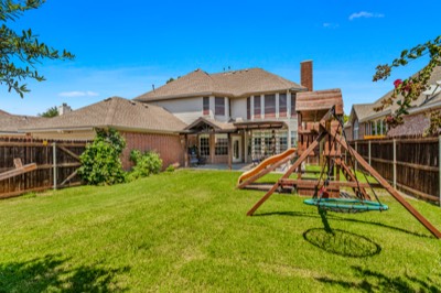 Grassy fenced backyard with wooden playset behind the brick home